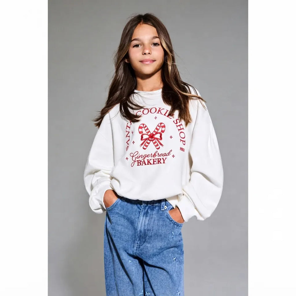 A young girl with long brown hair stands against a solid gray studio background wearing a white sweatshirt with a red 'Gingerbread Bakery' graphic and blue jeans.