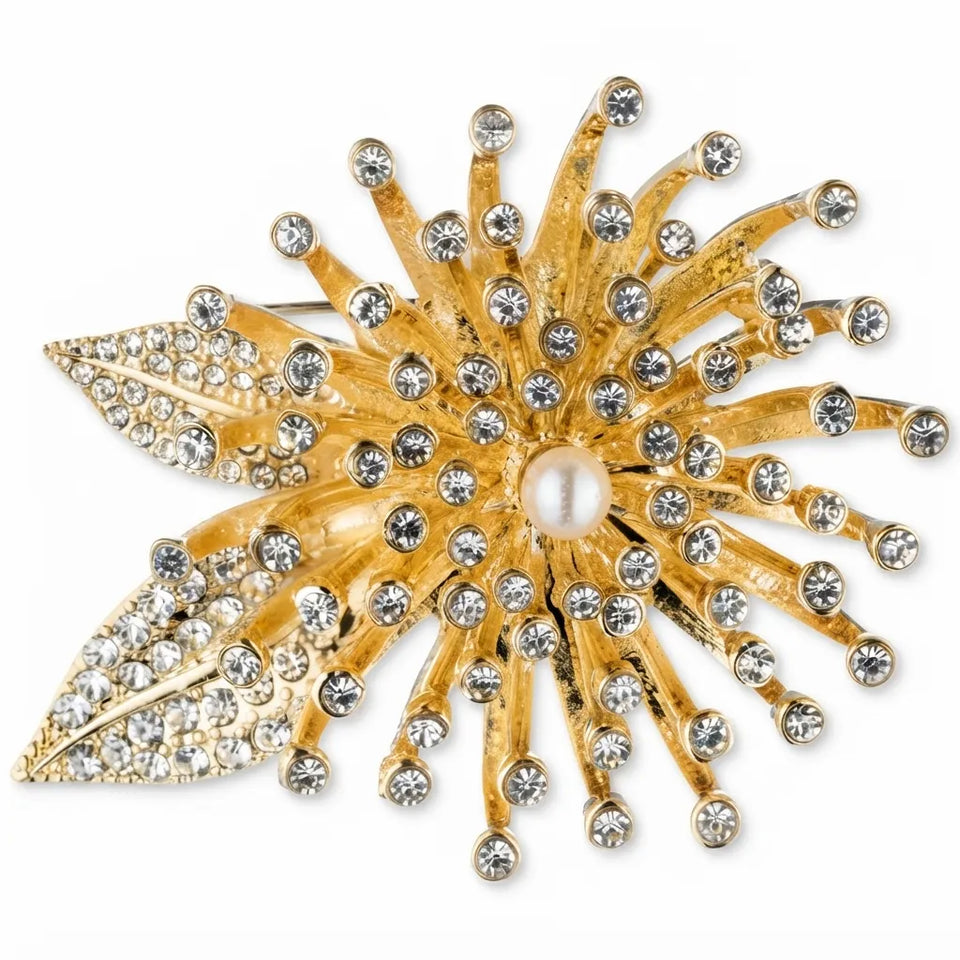 A gold-toned brooch shaped like a stylized flower with radiating leaf-like points, embellished with clear rhinestones and a central pearl, shown on a plain white background.