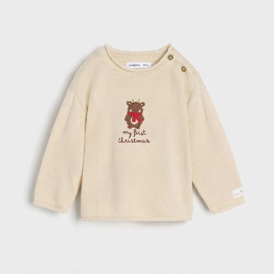 A cream-colored long-sleeve baby sweater with a teddy bear graphic wearing a red Santa hat and the text 'my first Christmas' on the chest. The top features two gold snap buttons on the shoulder.