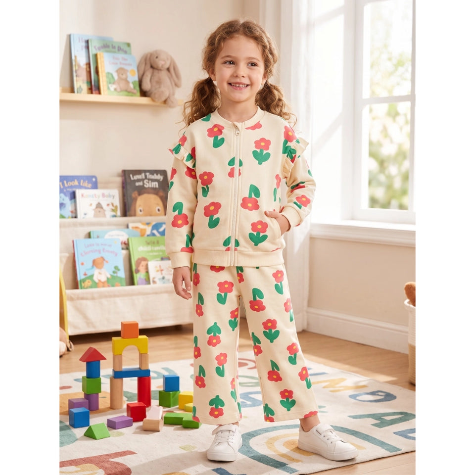 A young girl stands in a brightly lit children's room wearing a cream-colored zip-up sweatshirt and matching sweatpants set, both decorated with a colorful red and green floral pattern. She is smiling and posing on a patterned rug with toy blocks in front of her.