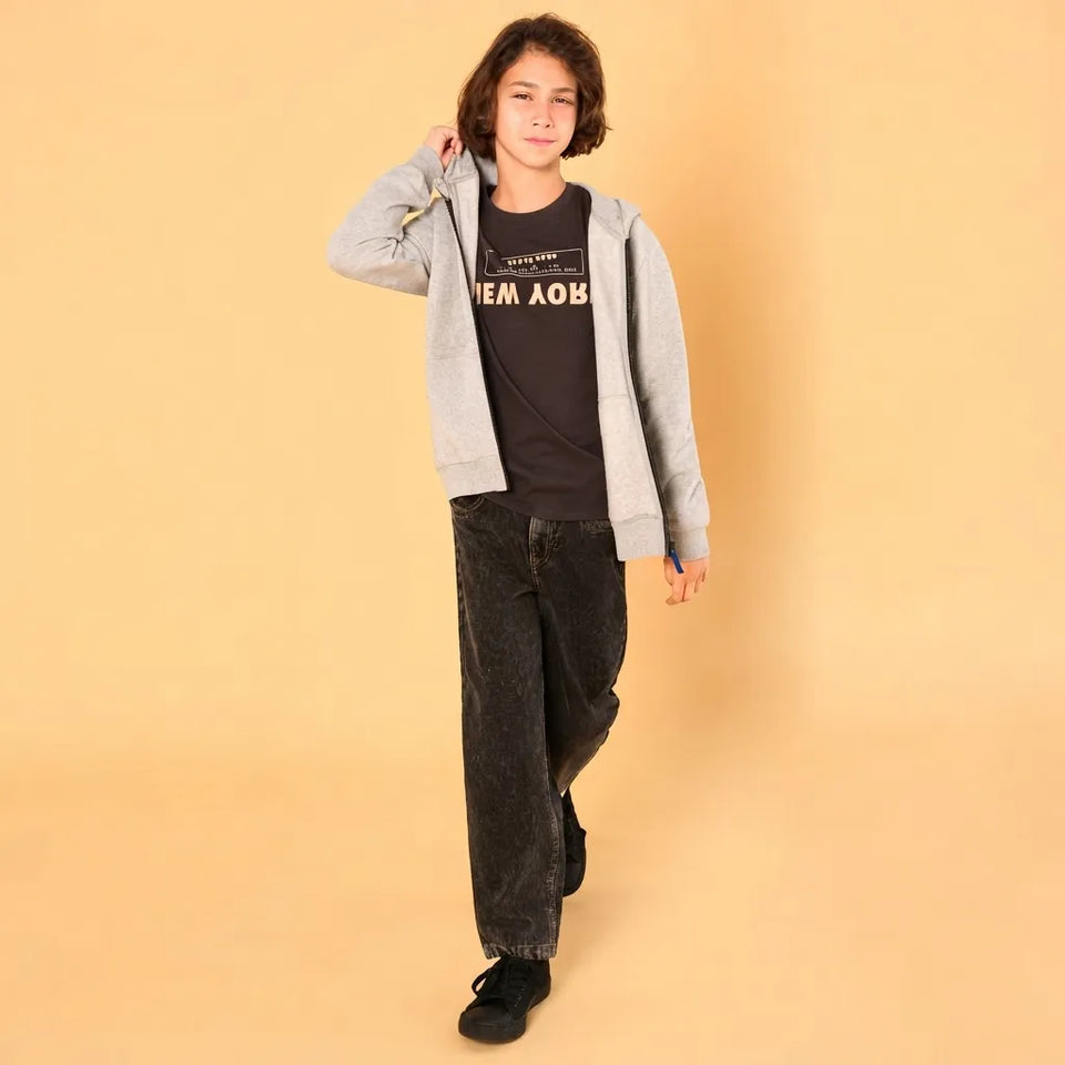 A young person modeling a light gray zip-up hoodie over a black graphic t-shirt, paired with dark gray washed jeans and black sneakers, against a solid beige background.