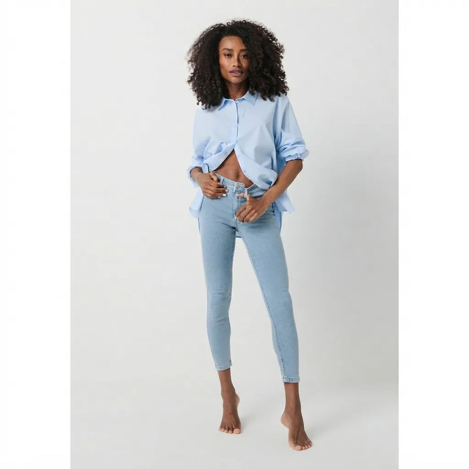 A woman modeling a light blue button-up shirt and matching light blue skinny jeans against a plain light gray studio background.