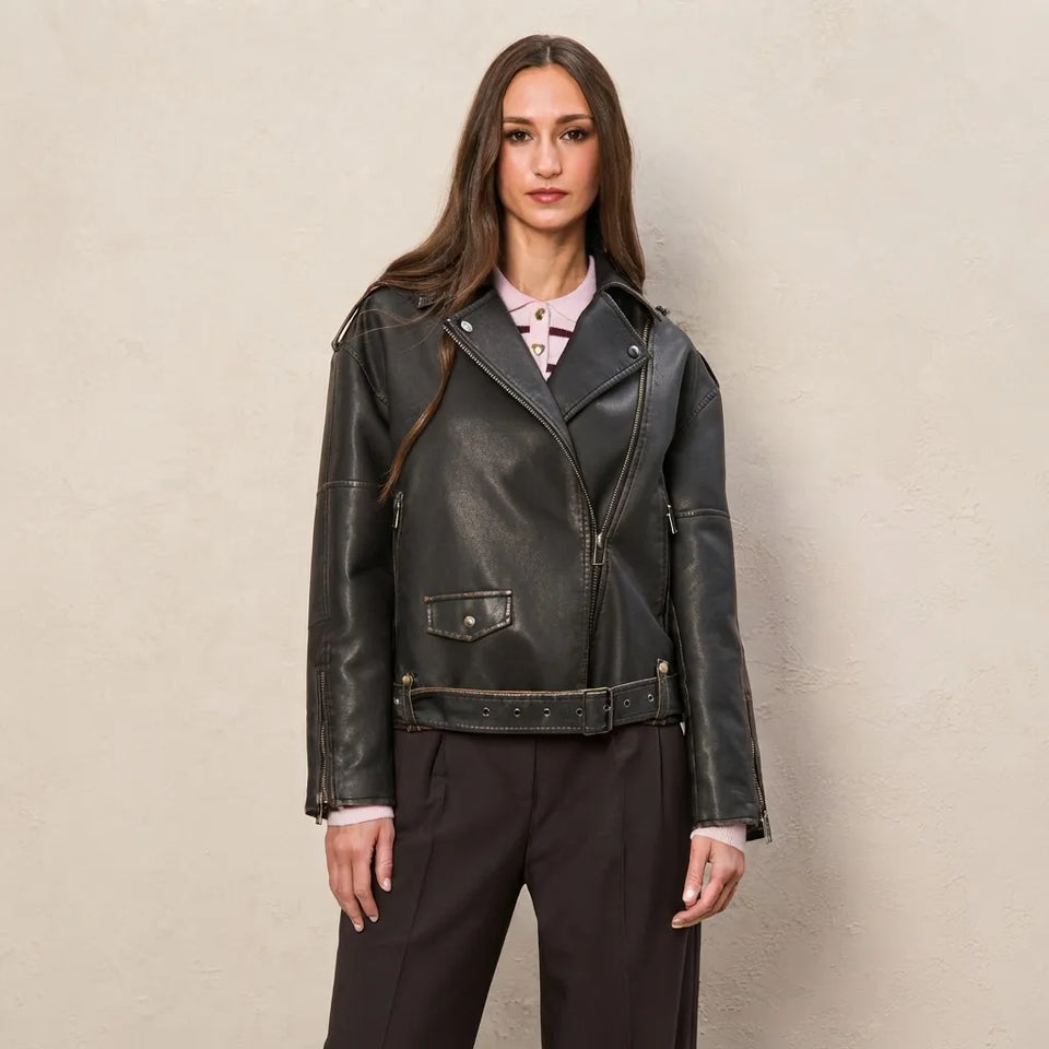 A woman modeling a black leather moto jacket with a zippered front, belted waist, and flap chest pocket, worn over a light pink collared shirt and dark trousers against a textured beige wall.