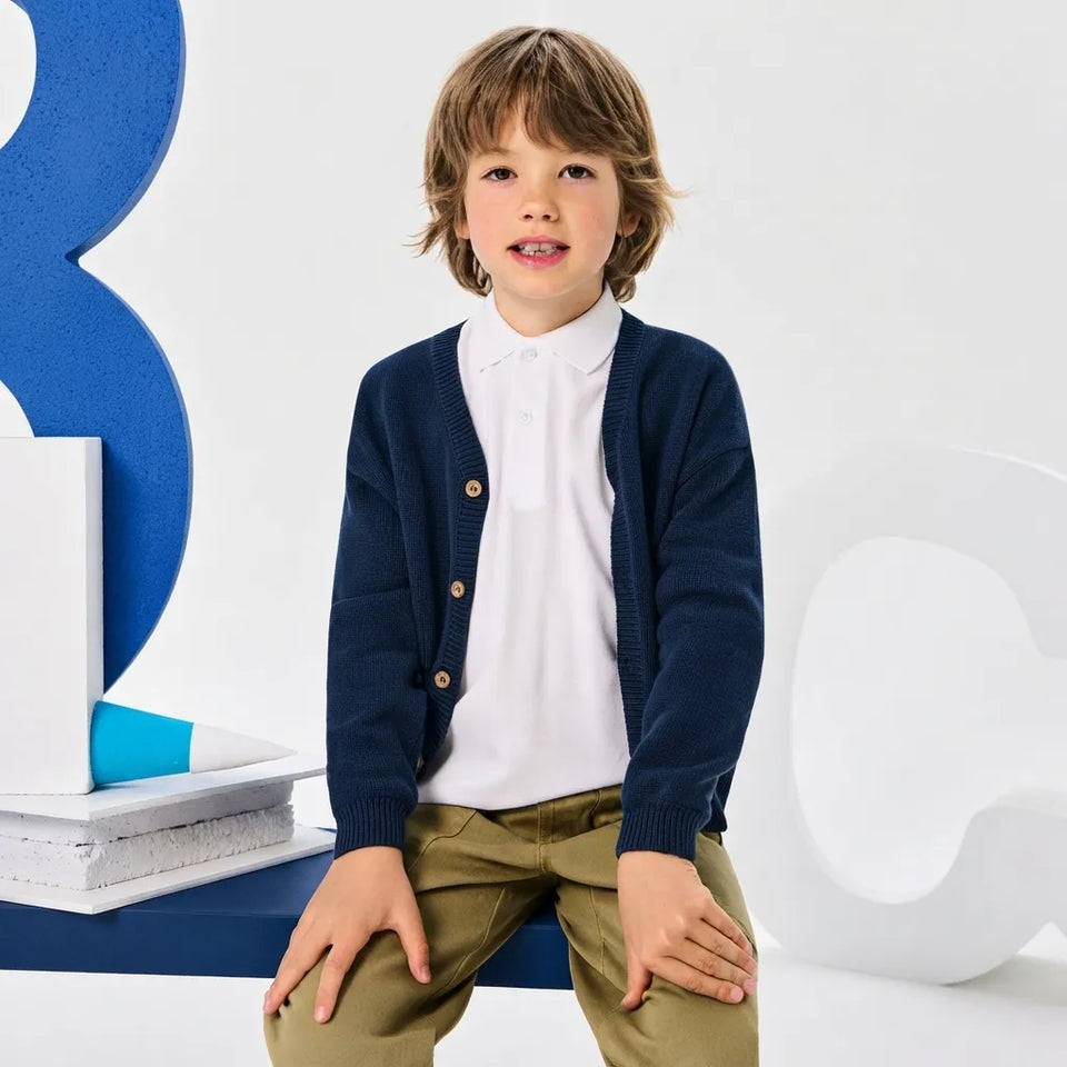A young boy wearing a navy blue knit cardigan over a white collared shirt and olive green trousers, sitting against a white studio background with large blue and white decorative letters.