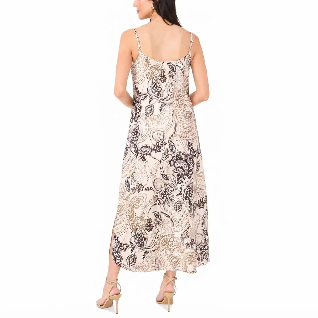 A woman modeling a sleeveless maxi dress from the back, featuring a beige base with a black and brown paisley pattern, paired with beige high-heeled sandals against a plain white background.