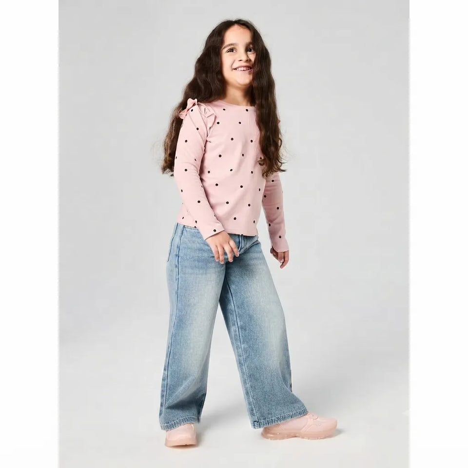 A young girl with long brown hair stands against a light gray studio background, wearing a light pink long-sleeve top with small black polka dots, light blue wide-leg jeans, and pale pink shoes.