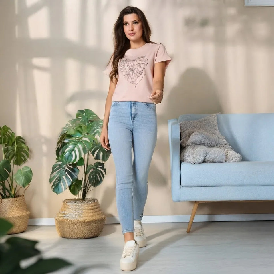 A woman modeling a light pink t-shirt with a sequined heart design, paired with light-wash blue skinny jeans and white sneakers, standing in a modern room with a light blue sofa and potted plants.