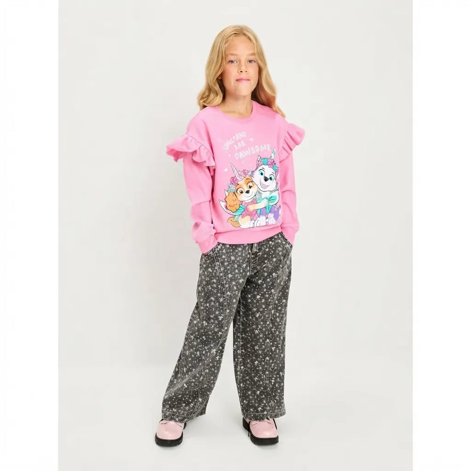 A young girl stands against a light gray studio background wearing a pink long-sleeve top with ruffled shoulders and a cartoon graphic on the front, paired with dark gray wide-leg pants covered in a small white floral pattern.