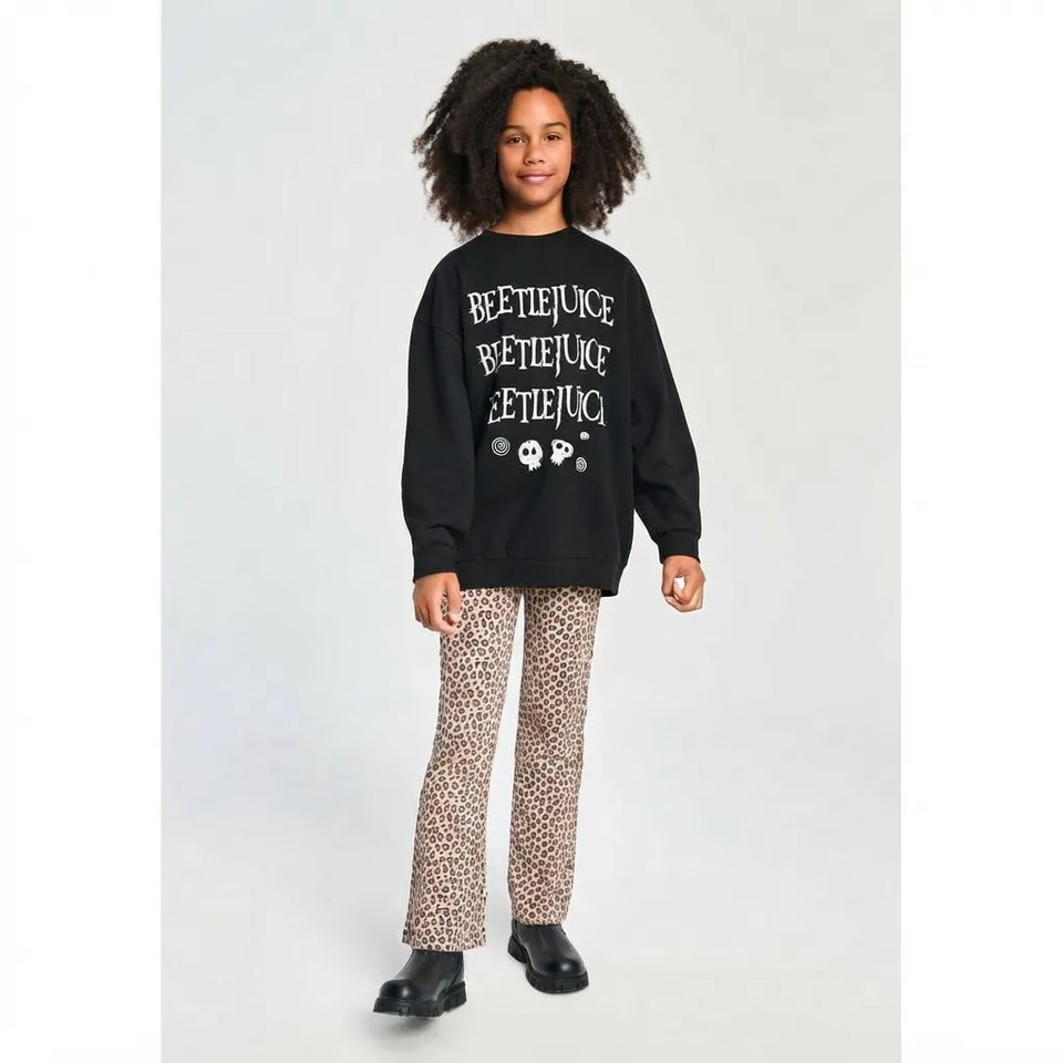 A child model wearing a black crewneck sweatshirt with white 'BEETJUICE' text and skull graphics, paired with beige and brown leopard-print pants and black shoes, standing against a light gray studio background.