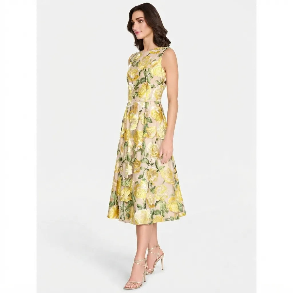 A woman modeling a sleeveless, knee-length A-line dress with a yellow and green floral print, paired with clear high-heeled sandals, against a light gray studio background.