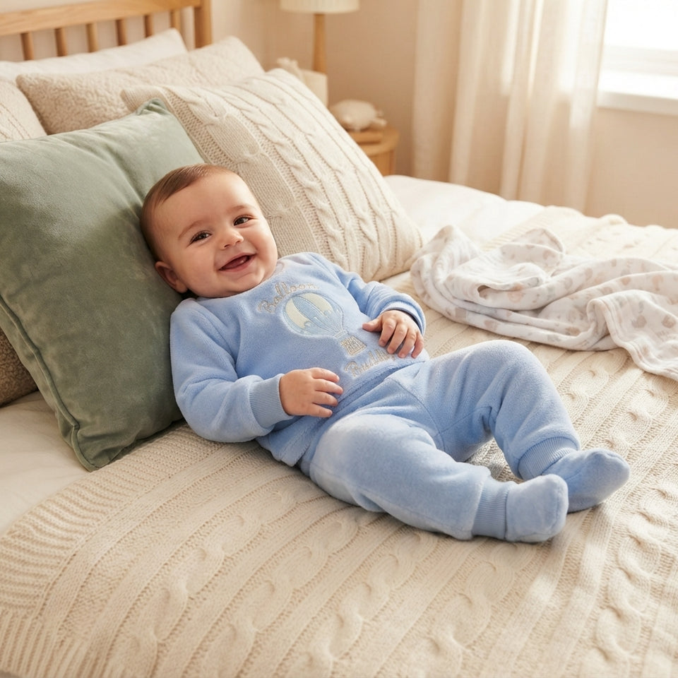 A smiling baby lying on a bed wearing a light blue footed one-piece outfit with a hot air balloon graphic. The scene is styled with cream-colored textured bedding, pillows, and a soft blanket in a bright, warm bedroom setting.