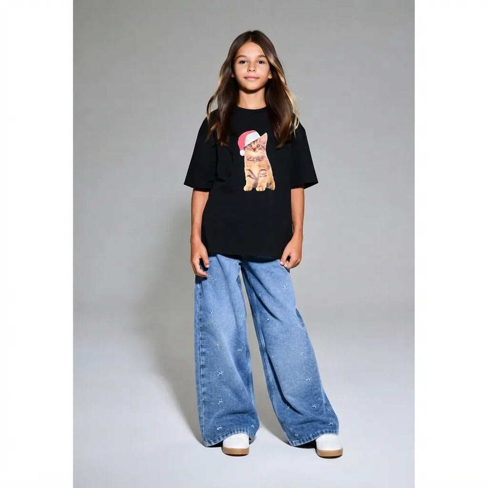 A young girl stands against a light gray studio background wearing a black graphic T-shirt with a cat illustration, wide-leg light blue jeans, and white sneakers.