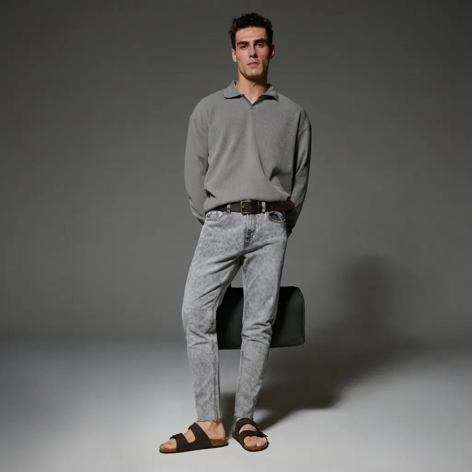 A man stands in a studio wearing a light gray long-sleeve polo shirt, light gray acid-wash jeans, a dark belt, and brown slide sandals. He is holding a black bag against a dark gray background.