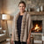 A woman wearing a beige knit poncho with a thick, fluffy faux-fur trim, standing in a cozy living room with a stone fireplace and a warm fire burning inside.