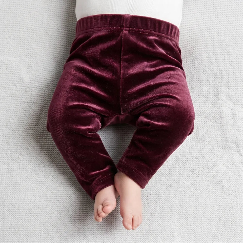 A close-up of a baby's lower body wearing plush, deep burgundy velvet pants, lying on a textured light gray blanket.