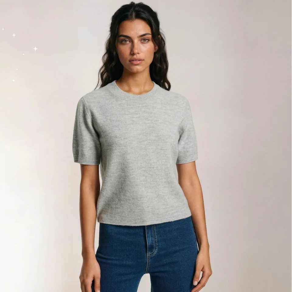 A woman modeling a light gray short-sleeve crewneck knit top with dark blue jeans, standing against a light beige studio background.