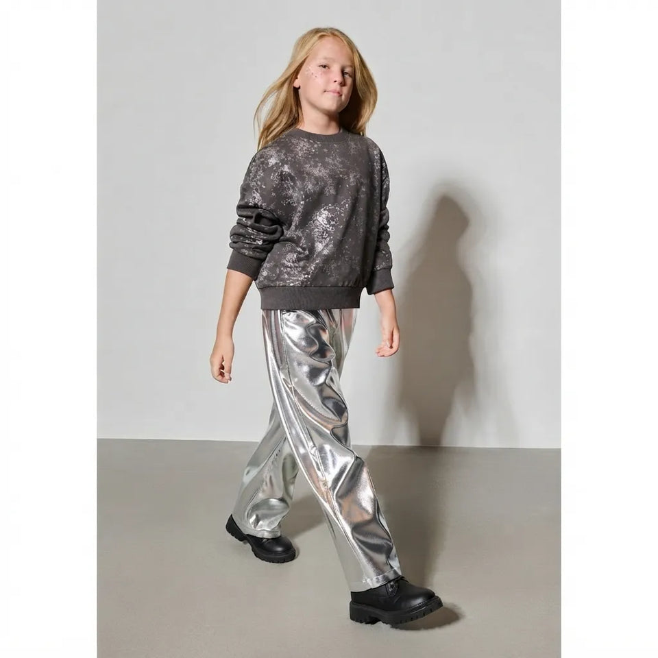 A young girl modeling a two-piece outfit consisting of a dark gray glittery sweatshirt and shiny metallic silver pants, paired with black boots, against a plain light gray studio background.