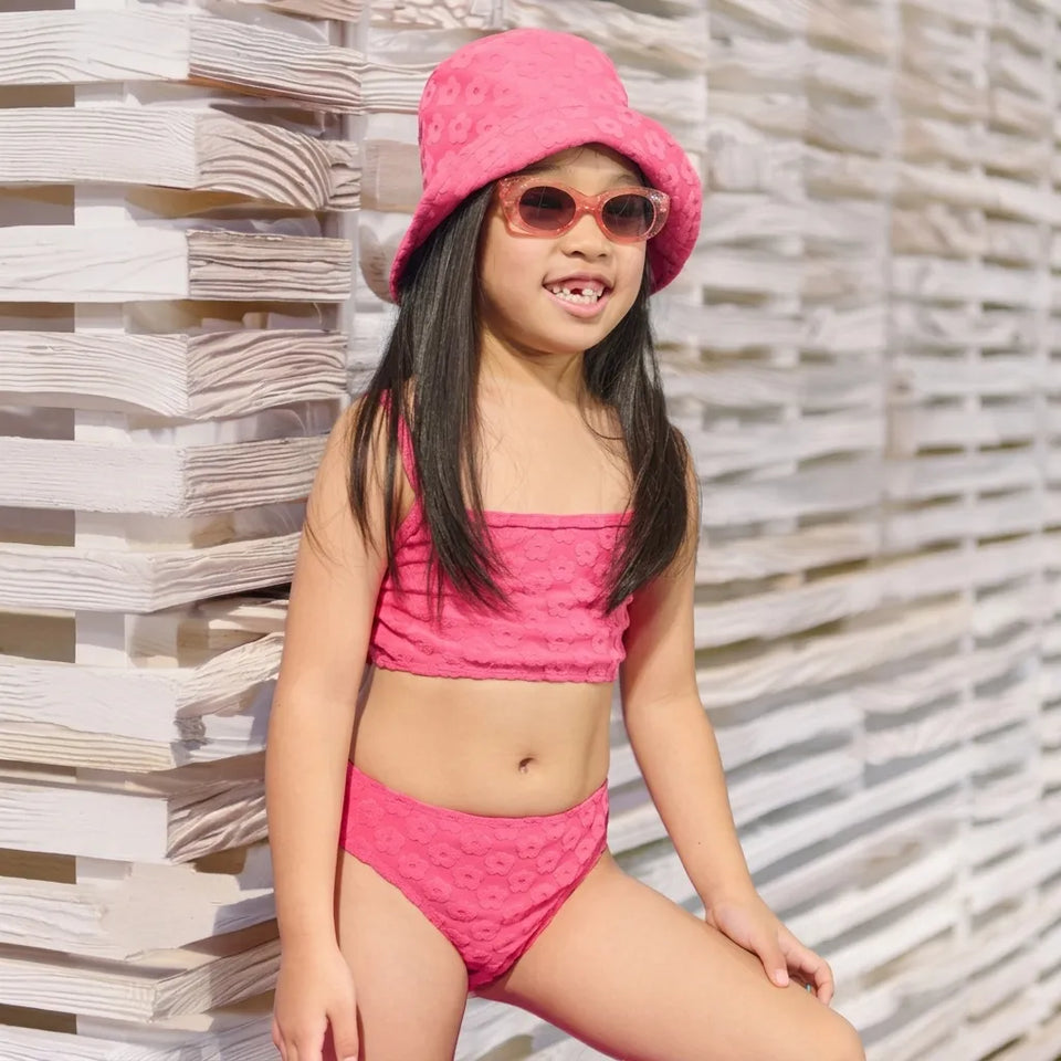 A young girl wearing a bright pink two-piece swimsuit with a matching pink textured bucket hat and pink sunglasses, posing in front of a white wooden slatted wall.