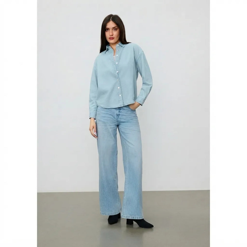 A woman modeling a light blue long-sleeve button-up shirt and matching light blue wide-leg jeans, standing against a neutral studio background.
