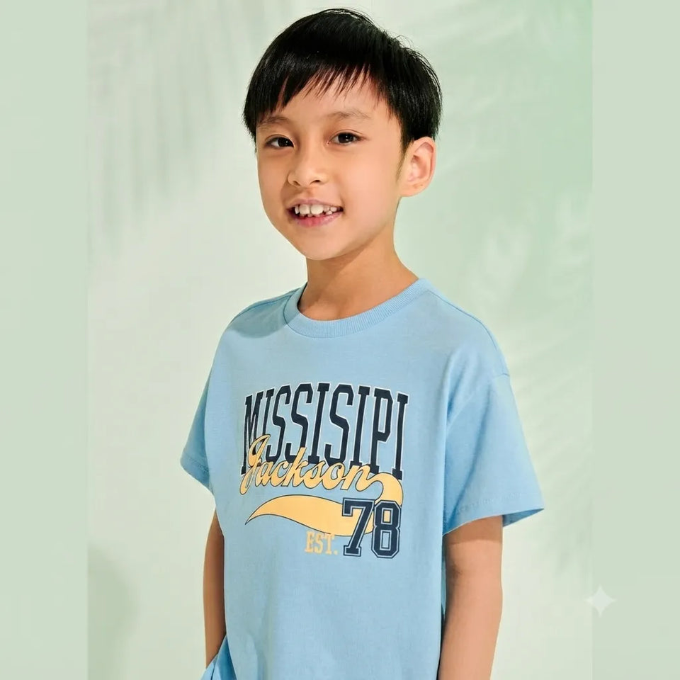 A young boy smiling and wearing a light blue t-shirt with 'MISSISSIPPI Jackson EST. 78' printed on the front, set against a soft, pale green background.
