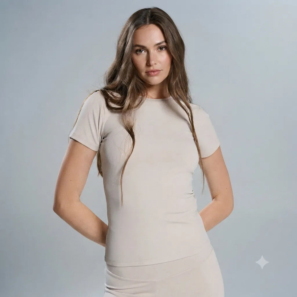 A woman modeling a fitted, short-sleeve beige t-shirt dress against a solid light gray studio background.
