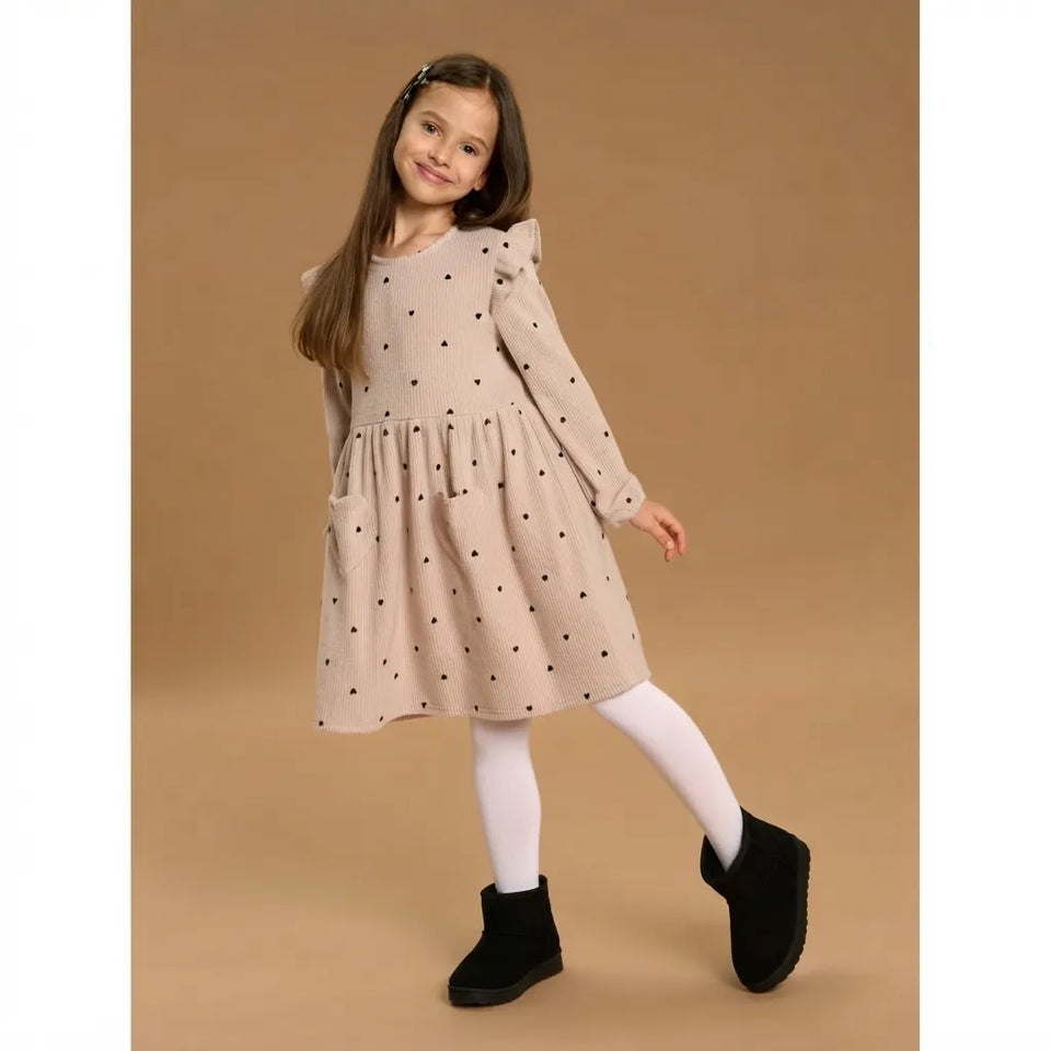 A young girl modeling a beige long-sleeve dress with black polka dots, white tights, and black boots, standing against a solid tan studio background.