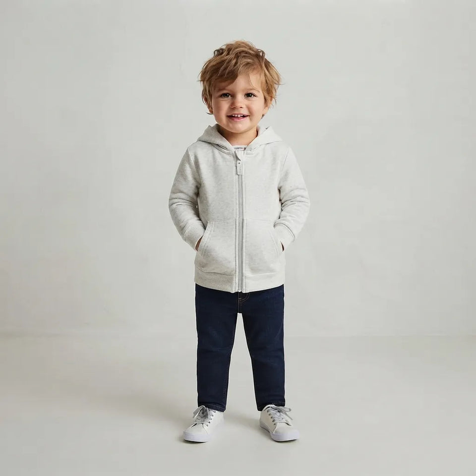 A toddler standing against a light gray studio background, wearing a light heather gray zip-up hoodie with a front pocket, navy pants, and white sneakers.