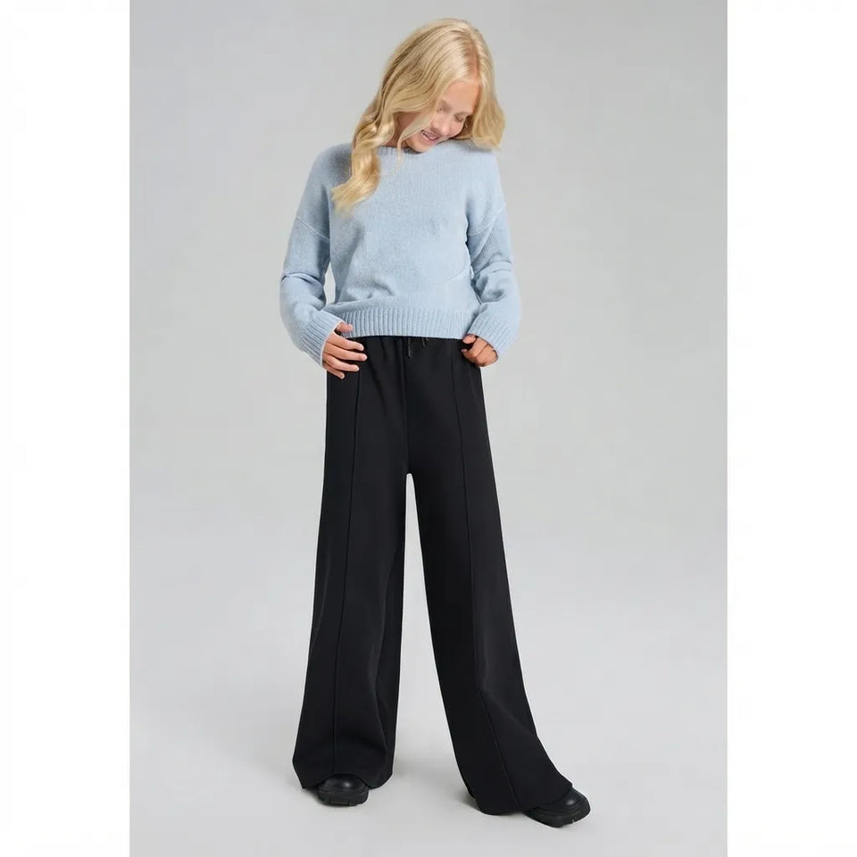 A young girl with blonde hair wearing a light blue knit sweater and black wide-leg trousers, standing against a neutral gray studio background.