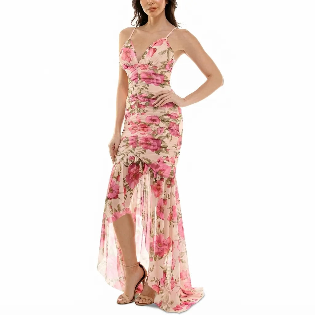 A woman modeling a sleeveless, high-low floral maxi dress with thin straps, a ruched bodice, and a sheer chiffon skirt with a pink and green floral print on a beige background.