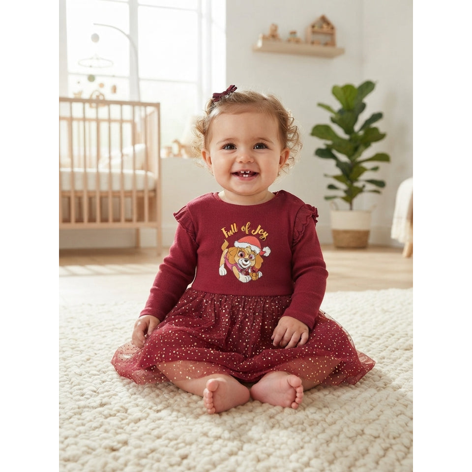 A baby girl sitting on a cream rug in a bright nursery, wearing a burgundy long-sleeve dress with a tulle skirt and a 'Haul of Joy' lion graphic on the front.