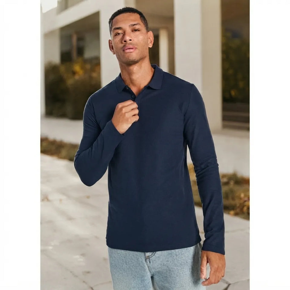 A man wearing a dark navy blue long-sleeve polo shirt and light gray jeans, standing outdoors in front of a modern building with light-colored columns.