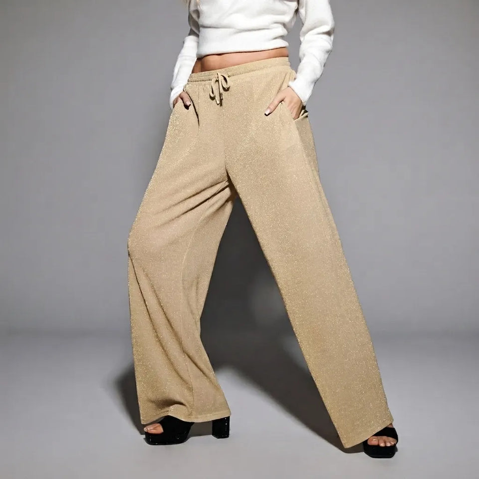 A model wearing wide-leg, knee-length pants in a textured beige fabric, styled with a white cropped long-sleeve top and black platform sandals against a gray studio background.