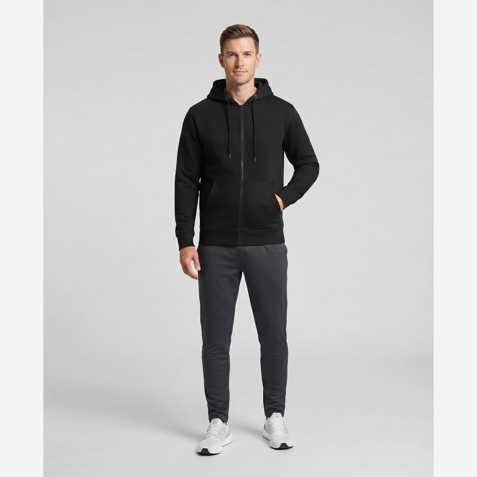 A man modeling a black zip-up hoodie with a drawstring hood and front pockets, paired with dark gray joggers and white sneakers, against a light gray studio background.