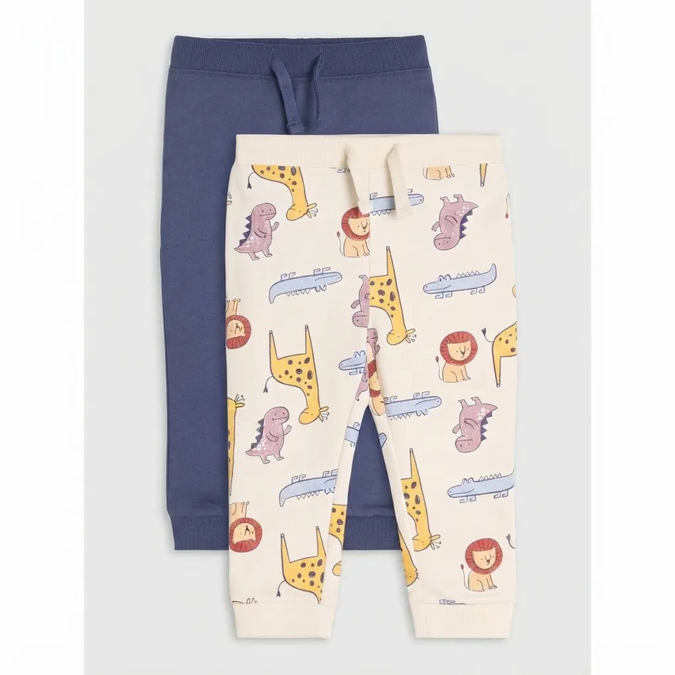 Two pairs of children's jogger pants laid flat on a light gray background. The front pair is cream with a colorful safari animal print including giraffes, lions, zebras, crocodiles, and airplanes.