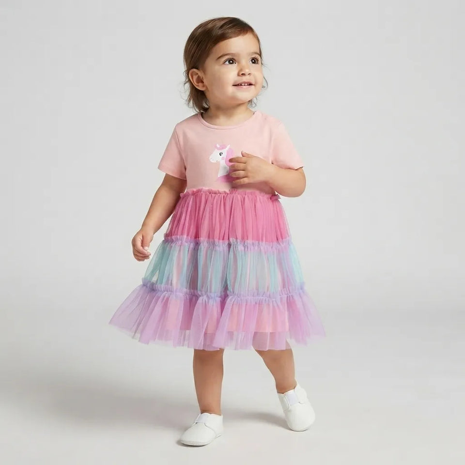 A toddler girl wearing a pink short-sleeve t-shirt with a unicorn graphic and a tiered tulle tutu skirt in pink, purple, and light blue, standing against a light gray studio background.