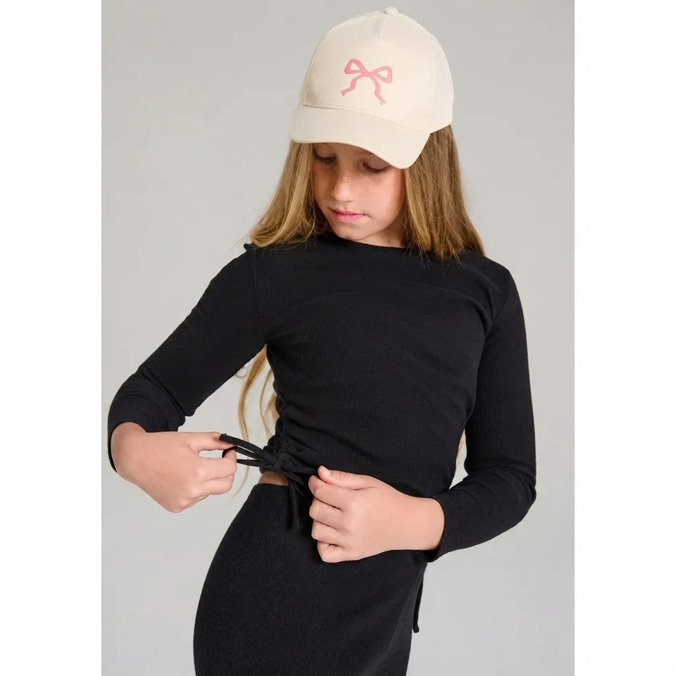 A young girl wearing a black long-sleeve ribbed top and matching black bottoms, with an off-white baseball cap featuring a pink bow. She is shown from the waist up, adjusting the tie at the waist of her top against a solid gray background.