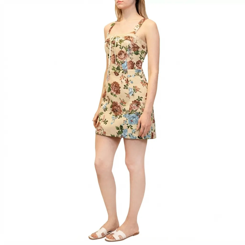 A woman modeling a sleeveless mini dress with a beige background and a colorful floral print, paired with light-colored slide sandals, against a plain white background.
