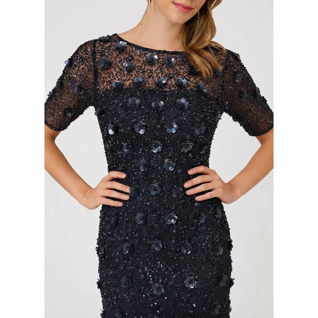 A woman wearing a black embellished evening gown with sheer lace short sleeves and sequined floral details, shown against a light gray studio background.