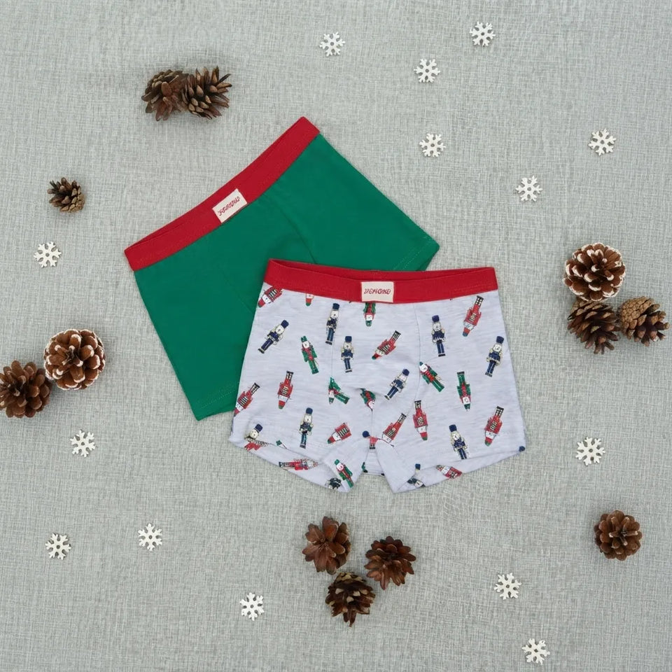 Two pairs of boxer briefs laid flat on a light gray fabric background with pinecones and white snowflake decorations. One pair is solid green with a red waistband, and the other is white with a red waistband and an all-over nutcracker print.