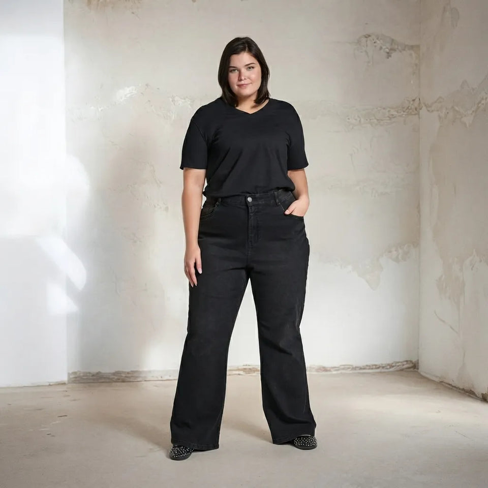 A woman modeling a black short-sleeve V-neck t-shirt and black wide-leg jeans, standing in a minimalist room with a textured off-white wall and concrete floor.