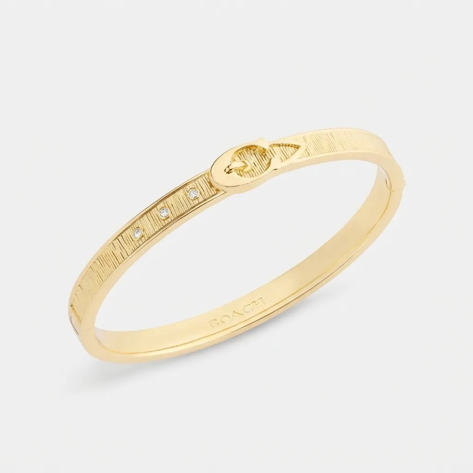 A gold bangle bracelet with a textured exterior, a small oval charm, and a row of tiny clear stones, shown on a light gray background.