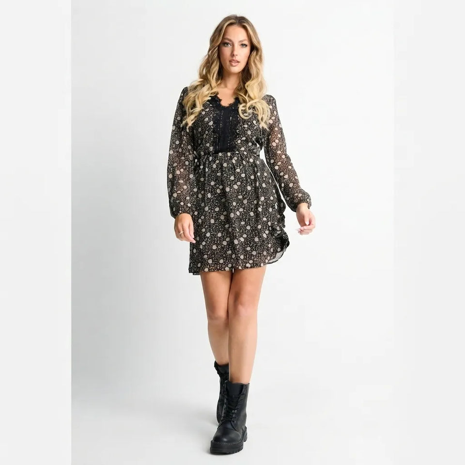 A woman modeling a black long-sleeve mini dress with a white floral pattern, featuring a smocked bodice and black combat boots, set against a plain light gray studio background.
