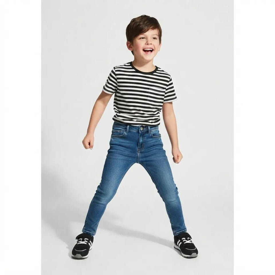 A young boy smiling and posing in a black and white striped short-sleeve t-shirt, blue skinny jeans, and black sneakers against a light gray studio background.