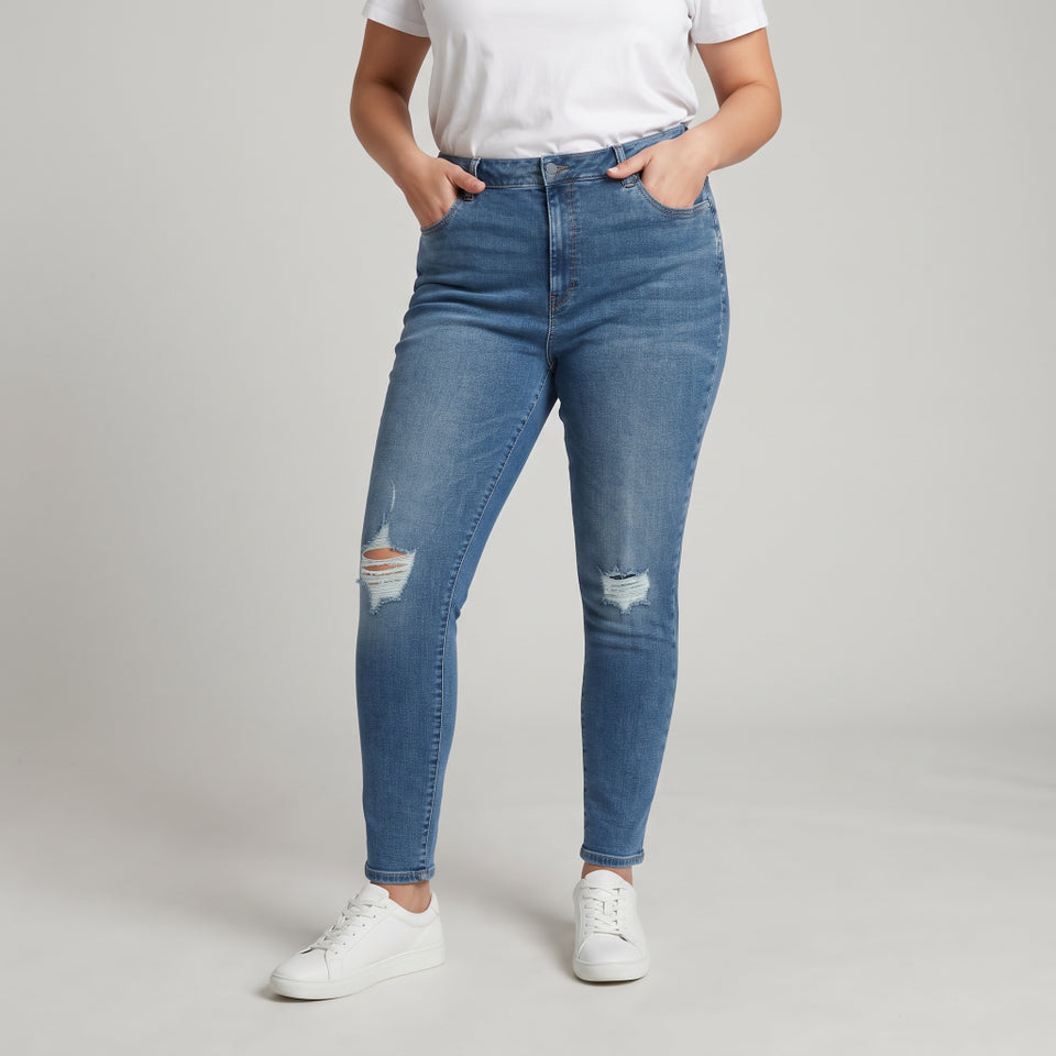 A model wearing medium-wash blue skinny jeans with distressed rips on the thighs and knees, paired with a white t-shirt and white sneakers, standing against a light gray studio background.