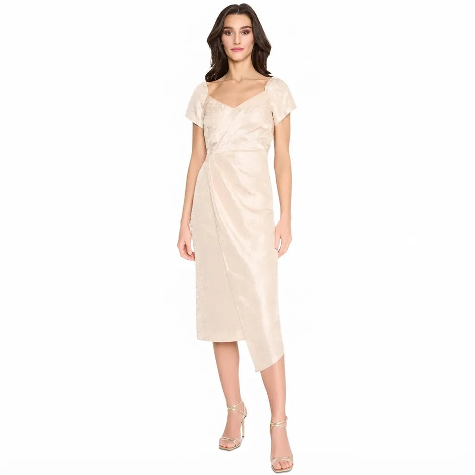 A woman modeling a champagne-colored midi wrap dress with short flutter sleeves and a draped waist, paired with clear high-heeled sandals, against a plain white background.