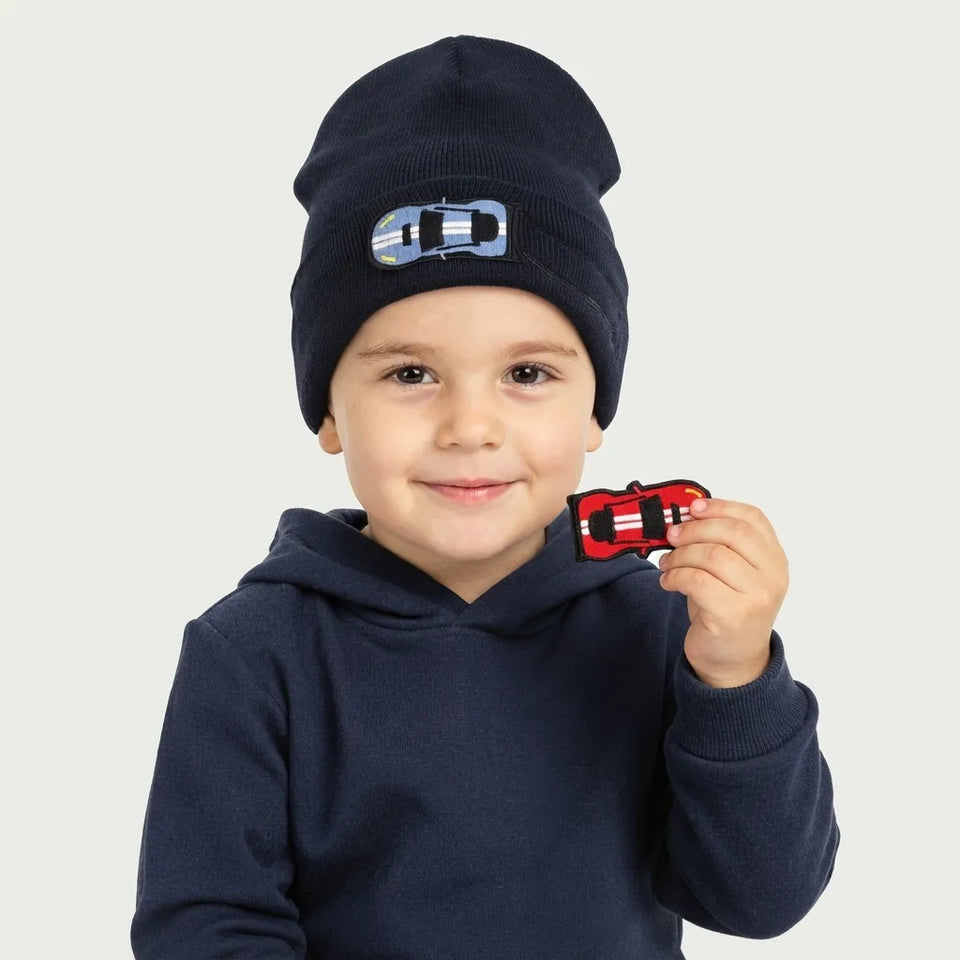 A young child wearing a navy blue knit beanie with a small car patch and holding a matching red car accessory, against a light gray background.