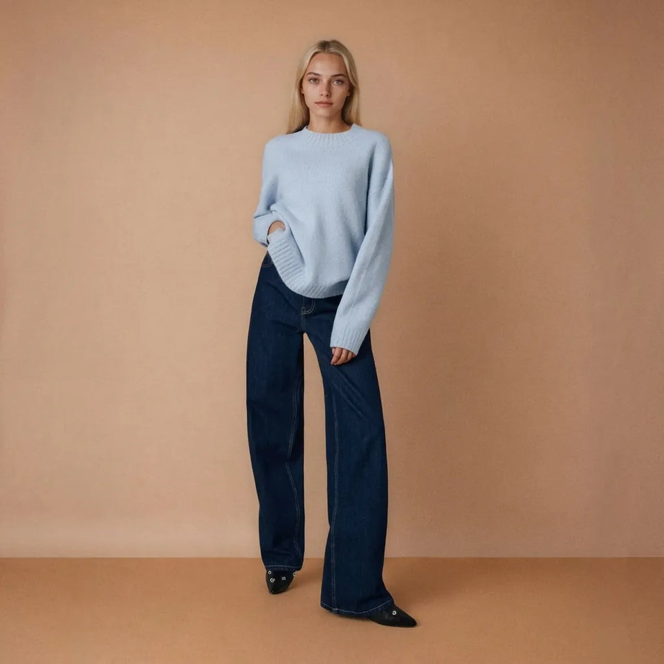 A woman stands against a warm beige studio backdrop wearing a light blue knit sweater and dark navy wide-leg trousers.