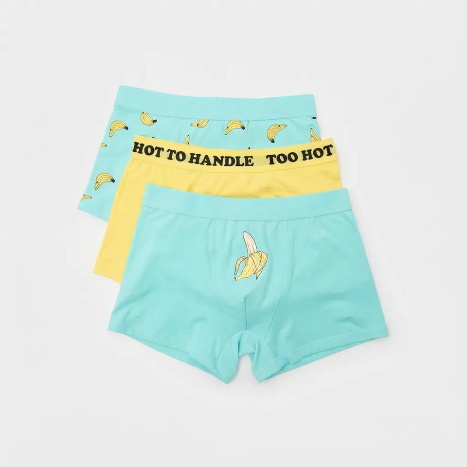 Three pairs of children's boxer briefs stacked on a light gray background. The top pair is light blue with a pattern of small yellow bananas and elastic waistband text reading 'HOT TO HANDLE TOO HOT'.
