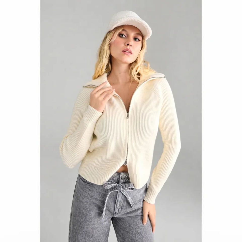 A woman modeling a cream ribbed knit zip-up hoodie with a half-zip collar, paired with gray jeans and a matching cream beanie, against a solid gray studio background.
