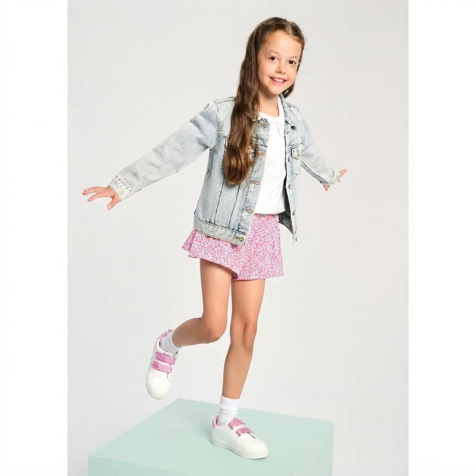 A young girl with long brown hair stands on a mint green pedestal, wearing a light-wash denim jacket, a white t-shirt, a pink floral mini skirt, and white sneakers with pink straps, against a light gray studio background.
